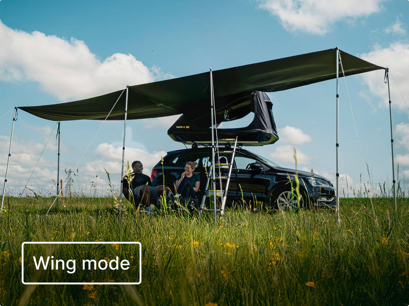 Two people sit by a car with the TentBox GO and Tunnel Awning set up in "wing mode".